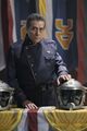 Commander William Adama at a table festooned with Colonial Fleet militaria during the service for the dead (TRS: "Miniseries, Night 2").