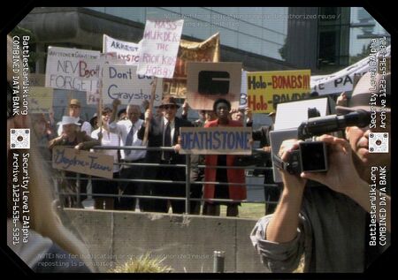 An unnamed woman dressed in a red coat holds a wide "Deathstone" sign amongst the other protesters behind a railed fence…