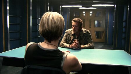 A room aboard Gemenon Traveler where Leoben Conoy is interrogated by Kara "Starbuck" Thrace, featuring windowed sliding…